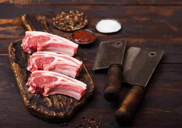 Carniceiros crus frescos costeletas de carne de cordeiro em tábua de corte com hatchets de carne vintage em fundo de madeira.Sal, pimenta e óleo em tigela de aço . — Fotografia de Stock