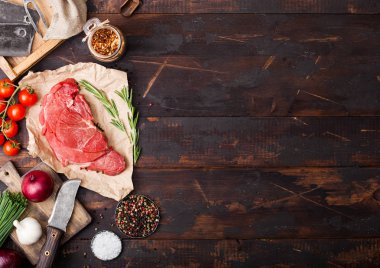 Fresh raw organic slice of braising steak fillet on butchers paper with fork and knife on dark wooden background. Red onion, tomatoes with salt and pepper. Space for text clipart