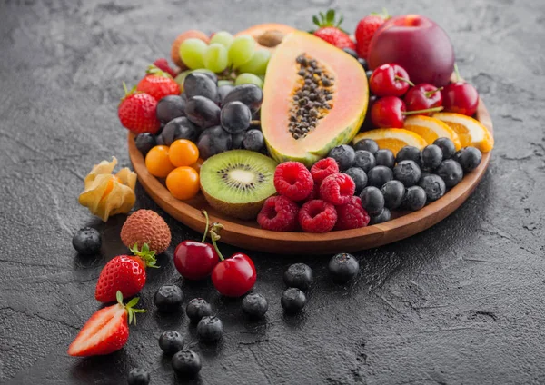 Fresh raw organic summer berries and exotic fruits in round wooden plate on black kitchen background. Papaya, grapes, nectarine, orange, raspberry, kiwi, strawberry, lychees, cherry and physalis. — Stock Photo, Image