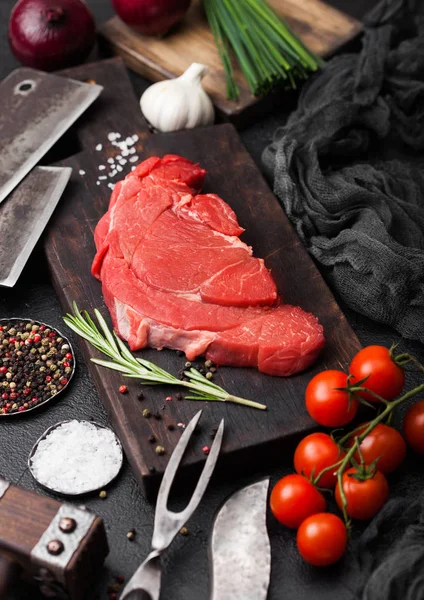 Fresh raw organic slice of braising steak fillet on chopping board with fork and knife on black stone background. Red onion, tomatoes with salt and pepper. — Stock Photo, Image