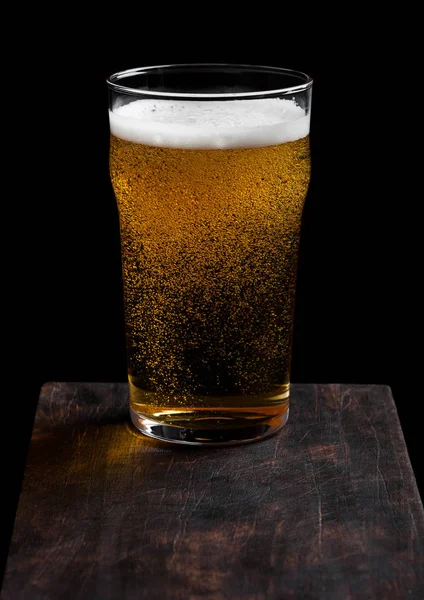 Glass of lager beer with foam and bubbles on vintage wooden board on black background. — Stock Photo, Image