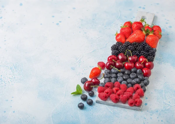 Fresh organic summer berries mix on white marble board on blue kitchen table background. Raspberries, strawberries, blueberries, blackberries and cherries. Space for text