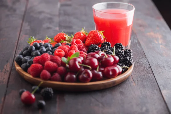 Fresh organic summer berries mix in round wooden tray with glass of juice on light wooden table background. Raspberries, strawberries, blueberries, blackberries and cherries.