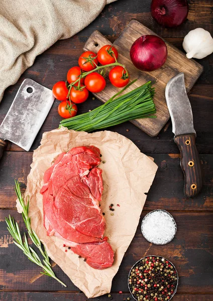 Čerstvý surový organický řez dušené bifteky na řezníky s vidličkou a nožem na tmavém dřevěném pozadí. Červená cibule, rajčata se solí a pepřem. — Stock fotografie