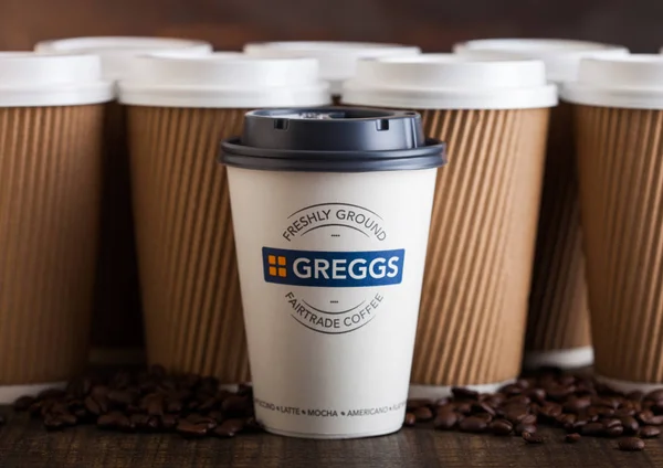 LONDRES, Reino Unido - 05 de junio de 2019: Greggs Coffee Paper Cup para llevar con granos de café sobre fondo de madera con tazas de café marrón en blanco . — Foto de Stock