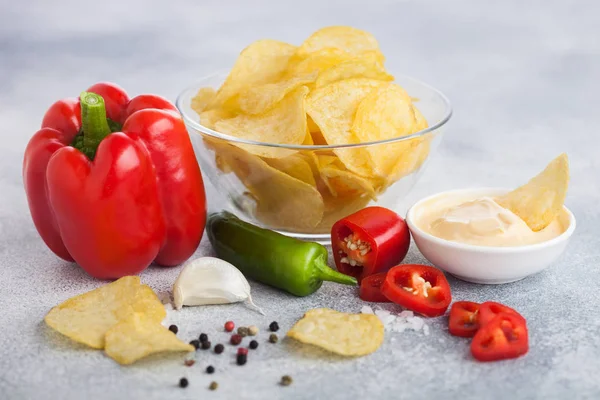 Glass bowl plate with potato crisps chips with onion flavour on light table background. Red and green chilli pepper with paprika and garlic. — Stock Photo, Image