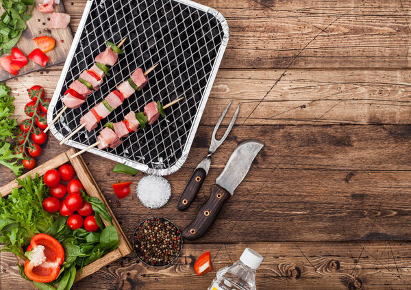Raw pork kebab with paprika on disposable coal bbq grill with fresh vegetables on wooden background with fork and knife. Salt and pepper with lettuce and paprika and cherry tomatoes.