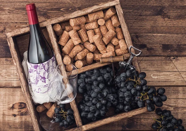 Bottle of red wine and empty glass with dark grapes with corks and opener inside vintage wooden box on grunge wooden background with linen towel. Top view