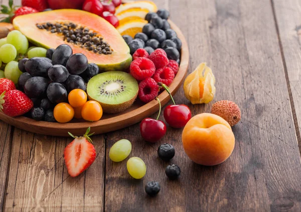 Fresh raw organic summer berries and exotic fruits in round wooden plate on wooden kitchen background. Papaya, grapes, nectarine, orange, raspberry, kiwi, strawberry, lychees, cherry. Macro — Stock Photo, Image