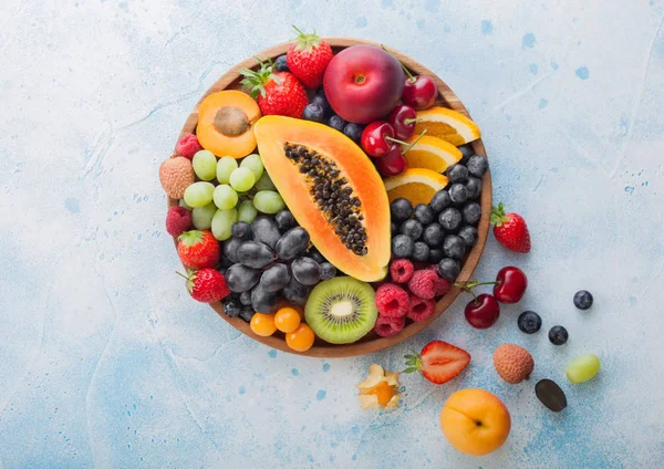 Frescas bayas de verano orgánicas crudas y frutas exóticas en plato redondo de madera sobre fondo de cocina azul claro. Papaya, uvas, nectarina, naranja, frambuesa, kiwi, fresa, lichis, cereza y physalis . — Foto de Stock