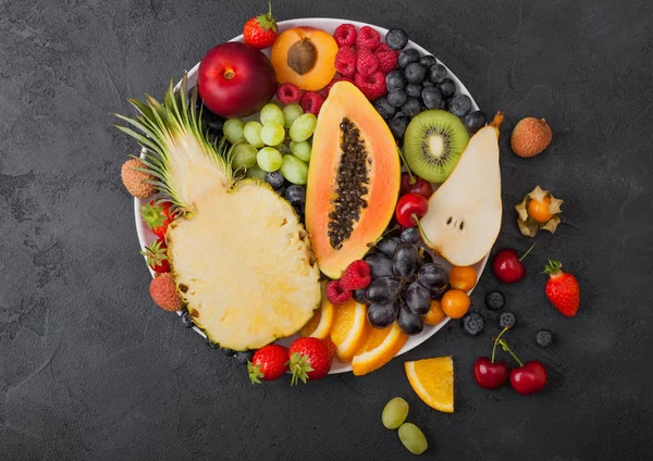 Bayas frescas de verano orgánicas crudas y frutas exóticas en plato blanco sobre fondo negro. Piña, papaya, uvas, nectarina, naranja, albaricoque, kiwi, pera, lichis, cereza y physalis . — Foto de Stock