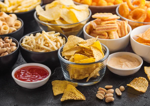 Todos los snacks clásicos de papa con cacahuetes, palomitas de maíz y aros de cebolla y pretzels salados en platos hondos sobre fondo negro. Giros con palitos y papas fritas y patatas fritas con nachos y bolas de queso . — Foto de Stock