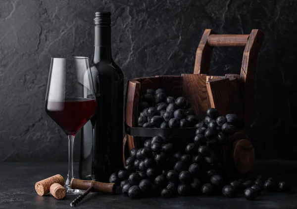 Elegant glass and bottle of red wine with dark grapes inside vintage wooden barrel on black stone background. Natural Light Stock Image