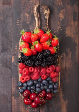 Koyu ahşap masa arka plan üzerinde vintage ahşap doğrama tahtası taze organik yaz meyveleri karışımı. Ahududu, çilek, yaban mersini, böğürtlen ve kiraz. 