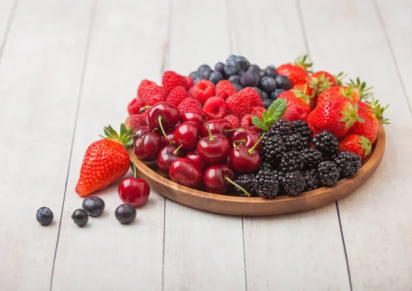 Fresh organic summer berries mix in round wooden tray on light wooden table background. Raspberries, strawberries, blueberries, blackberries and cherries.