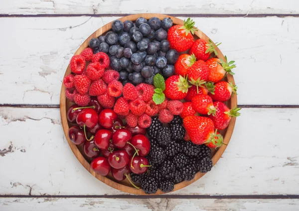 Las bayas frescas orgánicas veraniegas se mezclan en bandeja redonda de madera sobre fondo de mesa de madera blanca. Frambuesas, fresas, arándanos, moras y cerezas . — Foto de Stock