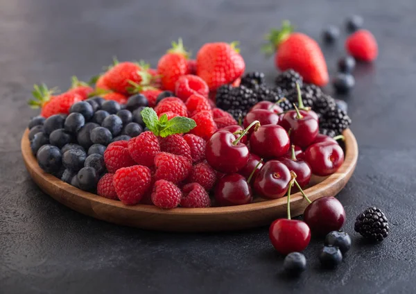 Fresh organic summer berries mix in round wooden tray on black kitchen table background. Raspberries, strawberries, blueberries, blackberries and cherries.