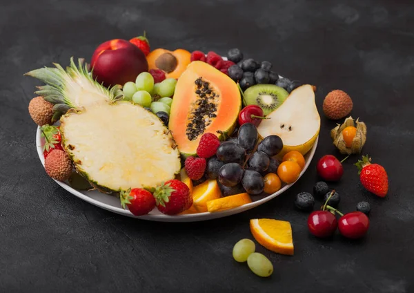 Bayas frescas de verano orgánicas crudas y frutas exóticas en plato blanco sobre fondo negro. Piña, papaya, uvas, nectarina, naranja, albaricoque, kiwi, pera, lichis, cereza y physalis . — Foto de Stock