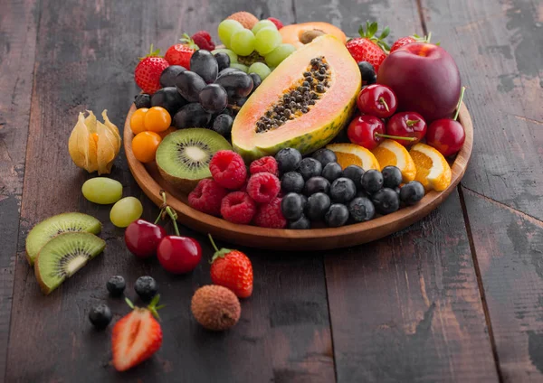 Fresh raw organic summer berries and exotic fruits in round wooden plate on dark wooden kitchen background. Papaya, grapes, nectarine, orange, raspberry, kiwi, strawberry, lychees, cherry. — Stock Photo, Image