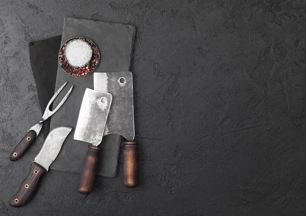 Coltello e forchetta da carne vintage e accette con tagliere in pietra e fondo tavola nero. Utensili da macellaio. Sale e pepe . — Foto Stock