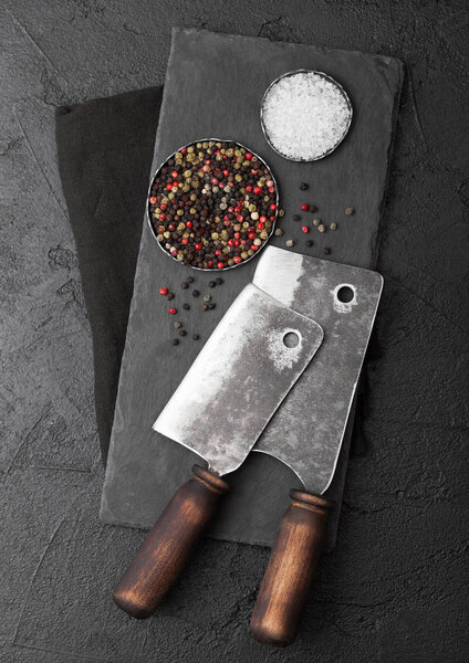 Vintage meat knife hatchets with stone chopping board and black table background. Butcher utensils. Salt and pepper. 