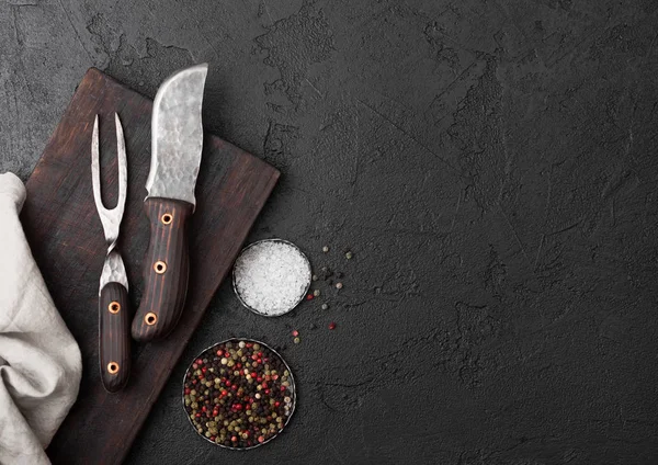 Vintage meat knife and fork on vintage chopping board and black stone table background. Butcher utensils. Salt and pepper. — Stock Photo, Image