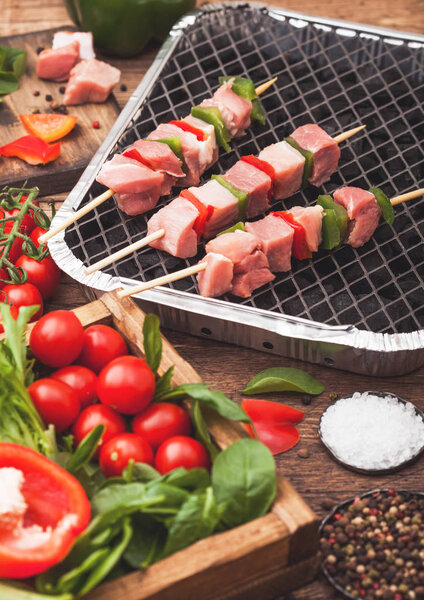 Сырая свинина кебаб с паприкой на одноразовом угольном барбекю гриль со свежими овощами на деревянном фоне с вилкой и ножом. Соль и перец с салатом и паприкой и помидорами черри. Вид сверху
