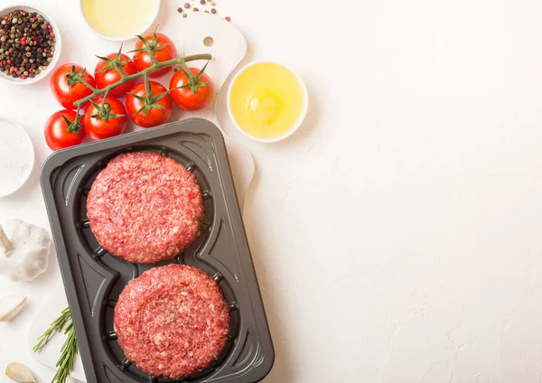 Plastic bakje met rauwe zelfgemaakte runderhamburgers met kruiden en specerijen. Bovenaanzicht en ruimte voor tekst boven op witte keuken tafel achtergrond met tomaten zout en peper. — Stockfoto