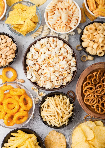 All classic potato snacks with peanuts, popcorn and onion rings and salted pretzels in bowl plates on light background. Twirls with sticks and potato chips and crisps with nachos and cheese balls.