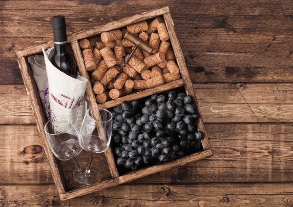 Bottle of red wine and empty glass with dark grapes with corks and opener inside vintage wooden box on grunge wooden background with linen towel. Top view