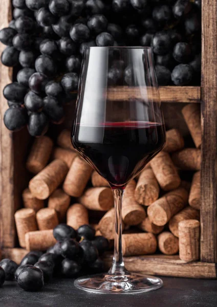 Copo elegante de vinho tinto com uvas escuras e rolhas dentro da caixa de madeira vintage em fundo de pedra preta. Luz natural — Fotografia de Stock