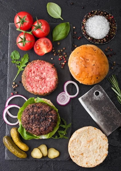 Hamburguesa de carne fresca a la parrilla y pimienta picada cruda en la tabla de cortar piedra con bollos de cebolla y tomates sobre fondo negro. Pepinillos salados y albahaca con hacha de carne . —  Fotos de Stock