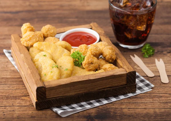 Buttered chicken nuggets and popcorn bites in vintage wooden box with ketchup and glass of cola on wooden background.