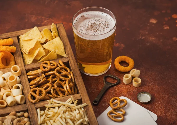 Glas ambachtelijk pils bier en opener met doos snacks op bruine achtergrond. Pretzel, zoute aardappelstokjes, pinda 's, uienringen met nacho' s in vintage doos met openers en biermatten. — Stockfoto