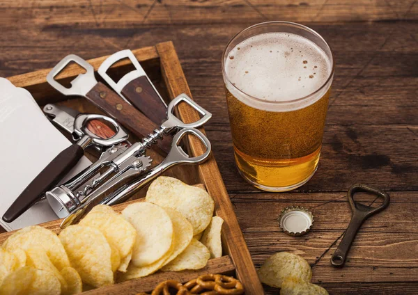 Glas ambachtelijk pils bier en opener met doos snacks op houten ondergrond. Pretzel en chips en zoute aardappelstokjes in vintage houten doos met openers en biermatten. Blauwe wijting — Stockfoto