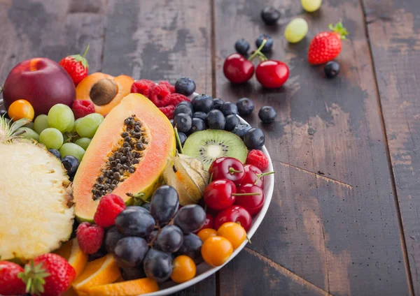 Fresh raw organic summer berries and exotic fruits in white plate on dark wooden background. Pineapple, papaya, grapes, nectarine, orange, apricot, kiwi, strawberry, lychees, cherry. Macro — Stock Photo, Image