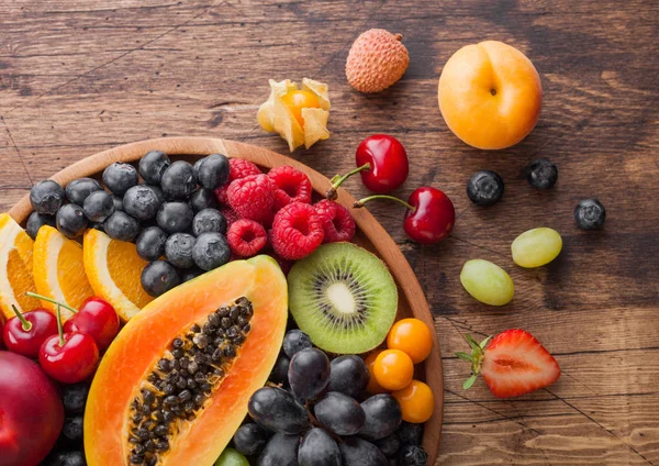 Свежие органические летние ягоды и экзотические фрукты в круглой деревянной тарелке на деревянном кухонном фоне. Папайя, виноград, нектарин, апельсин, малина, киви, клубника, лишайник, вишня. Макро — стоковое фото
