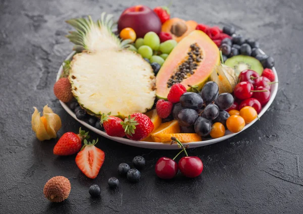 Des baies fraîches d'été bio crues et des fruits exotiques dans une assiette blanche sur fond noir. Ananas, papaye, raisin, nectarine, orange, abricot, kiwi, poire, litchi, cerise et physalis . — Photo