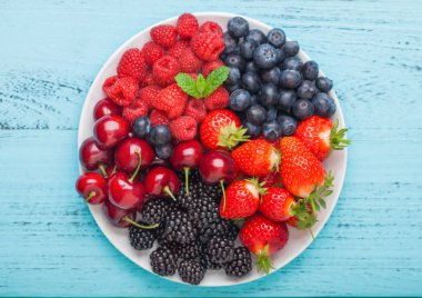 Taze organik yaz meyveleri mavi ahşap masa arka plan üzerinde beyaz plaka karışımı. Ahududu, çilek, yaban mersini, böğürtlen ve kiraz. 