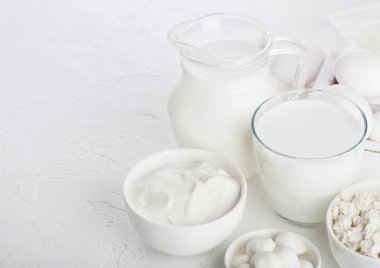 Beyaz masa arka planında taze süt ürünleri. Kavanoz ve bir bardak süt, bir kase ekşi krema, süzme peynir ve mozzarella. Tahta kutuda yumurtalar. Metin için boşluk