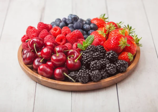 Φρέσκα βιολογικά φρούτα του καλοκαιριού αναμειγνύονται σε στρογγυλό ξύλινο δίσκο σε ανοιχτόχρωμο ξύλινο φόντο τραπεζιού. Σμέουρα, φράουλες, βατόμουρα, βατόμουρα και κεράσια. — Φωτογραφία Αρχείου