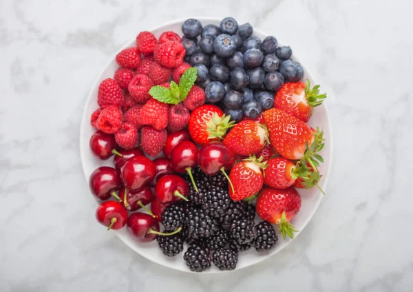 Fresh organic summer berries mix in white plate on marble background. Raspberries, strawberries, blueberries, blackberries and cherries.