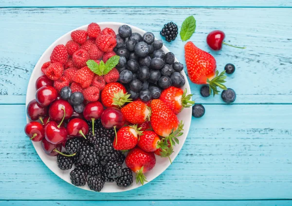Les baies fraîches d'été bio se mélangent dans une assiette blanche sur fond de table en bois bleu. Framboises, fraises, bleuets, mûres et cerises . — Photo