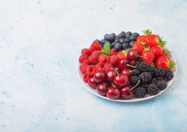 Les baies d'été bio fraîches se mélangent dans une assiette blanche sur fond bleu de table de cuisine. Framboises, fraises, bleuets, mûres et cerises. Espace pour le texte — Photo