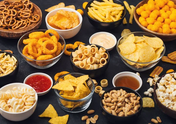 Todos los snacks clásicos de papa con cacahuetes, palomitas de maíz y aros de cebolla y pretzels salados en platos hondos sobre fondo negro. Giros con palitos y papas fritas y patatas fritas con nachos y bolas de queso . — Foto de Stock