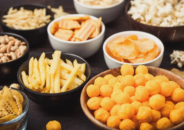 All classic potato snacks with peanuts, popcorn and onion rings and salted pretzels in bowl plates on wooden background. Twirls with sticks and potato chips and crisps with nachos and cheese balls. — Stock Photo, Image