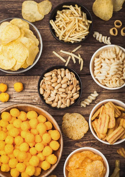 Todos los snacks clásicos de papa con cacahuetes, palomitas de maíz y aros de cebolla y pretzels salados en platos hondos sobre fondo de madera. Giros con palitos y papas fritas y patatas fritas con nachos y bolas de queso . — Foto de Stock