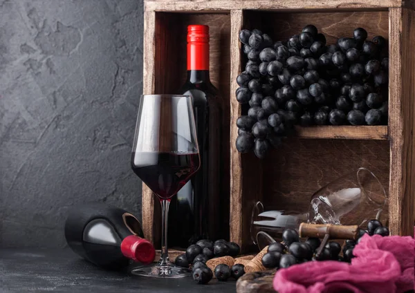 Garrafas de vinho tinto com uvas escuras dentro da caixa de madeira vintage em fundo de pedra preta. Copo de vinho elegante com rolhas e saca-rolhas em placa preta.Luz natural — Fotografia de Stock