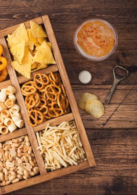 Bir bardak bira ve ahşap arka planda atıştırmalık kutusu. Pretzel, tuzlu patates çubukları, fıstık, nachos 'lu soğan halkaları, açıcılar ve bira paspasları. Üst görünüm
