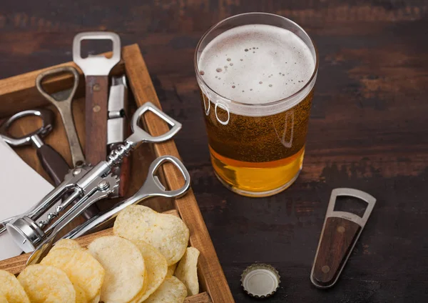 Glas ambachtelijk pils bier en opener met doos snacks op houten ondergrond. Pretzel en chips en zoute aardappelstokjes in vintage houten doos met openers en biermatten. Blauwe wijting — Stockfoto